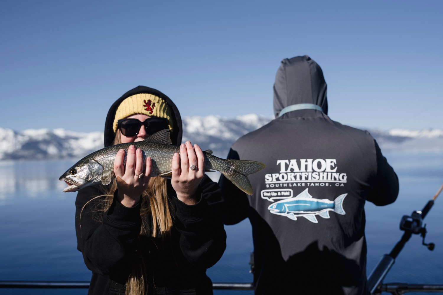 Person holding a fish with snowy mountains and a lake in the background, while another person stands beside them.