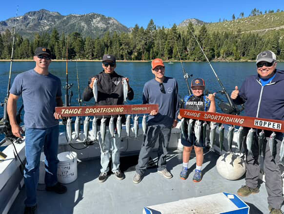 Five people holding fish on a boat with mountains and trees in the background.