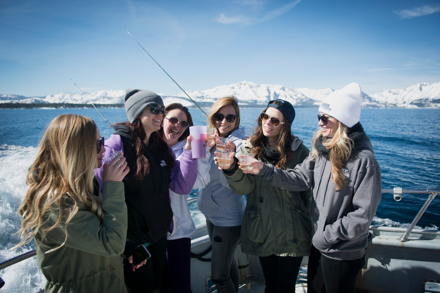 Six women on a boat holding drinks, with snowy mountains in the background.