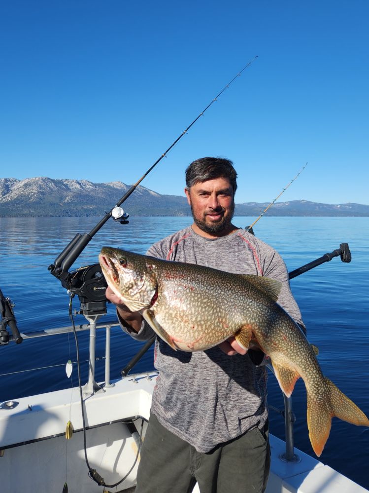 Man on a boat holding a large fish with two fishing rods and scenic mountains in the background.