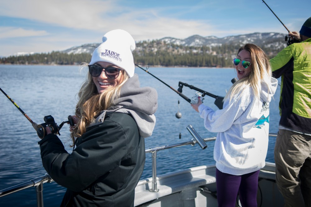 Two people smiling while fishing on a boat, with mountains in the background and a clear blue sky.