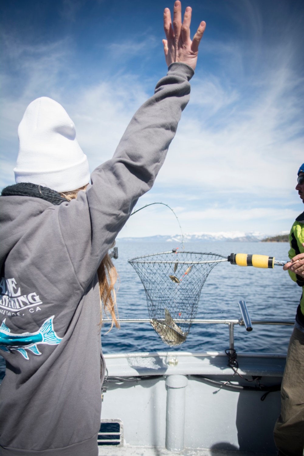 Person on boat raises arm while holding a fishing net with a fish over a lake.