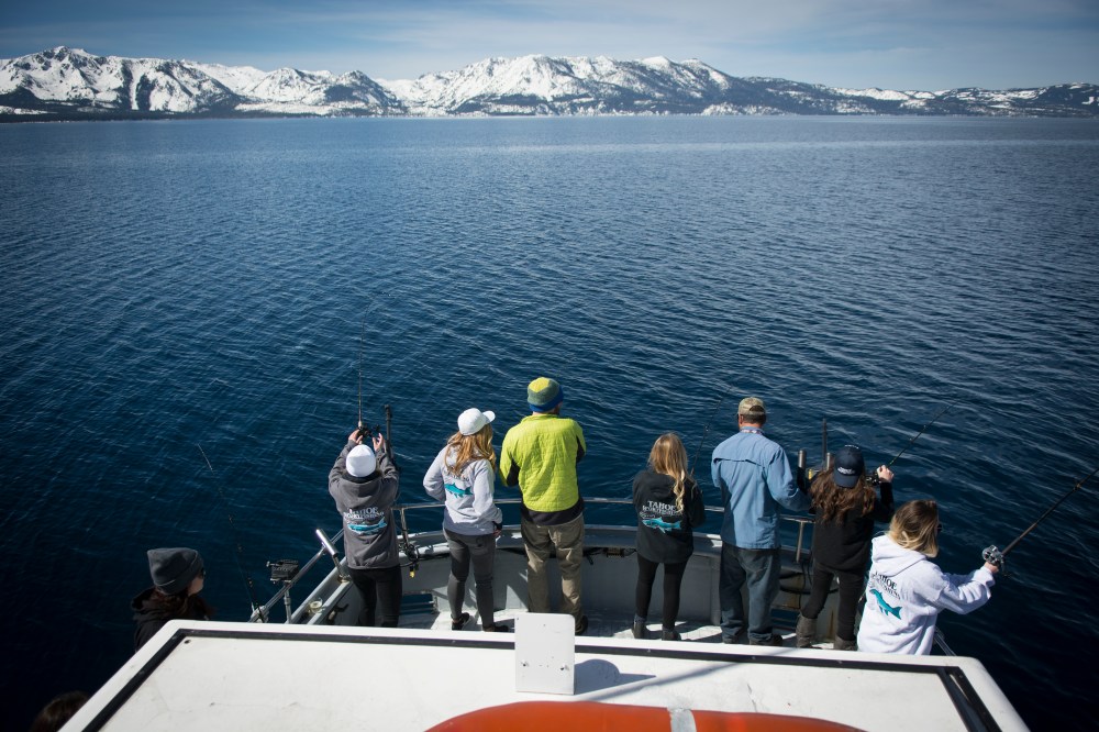Group of people fishing from a boat on a lake with snowy mountains in the background.