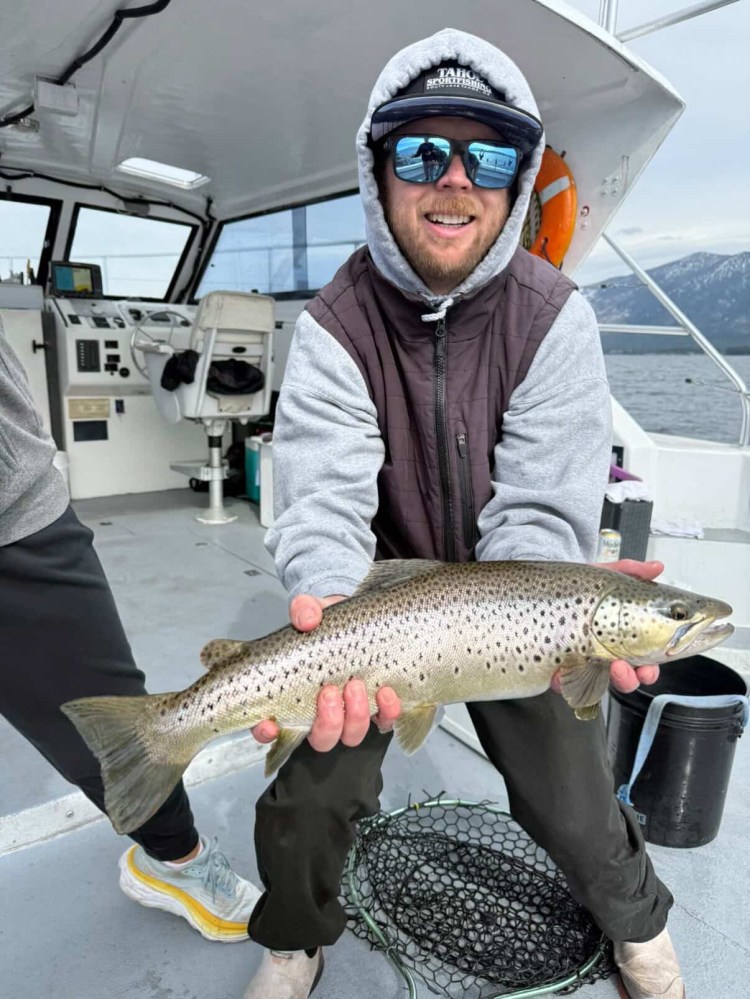 Person in sunglasses holding a large fish on a boat with mountains in the background.