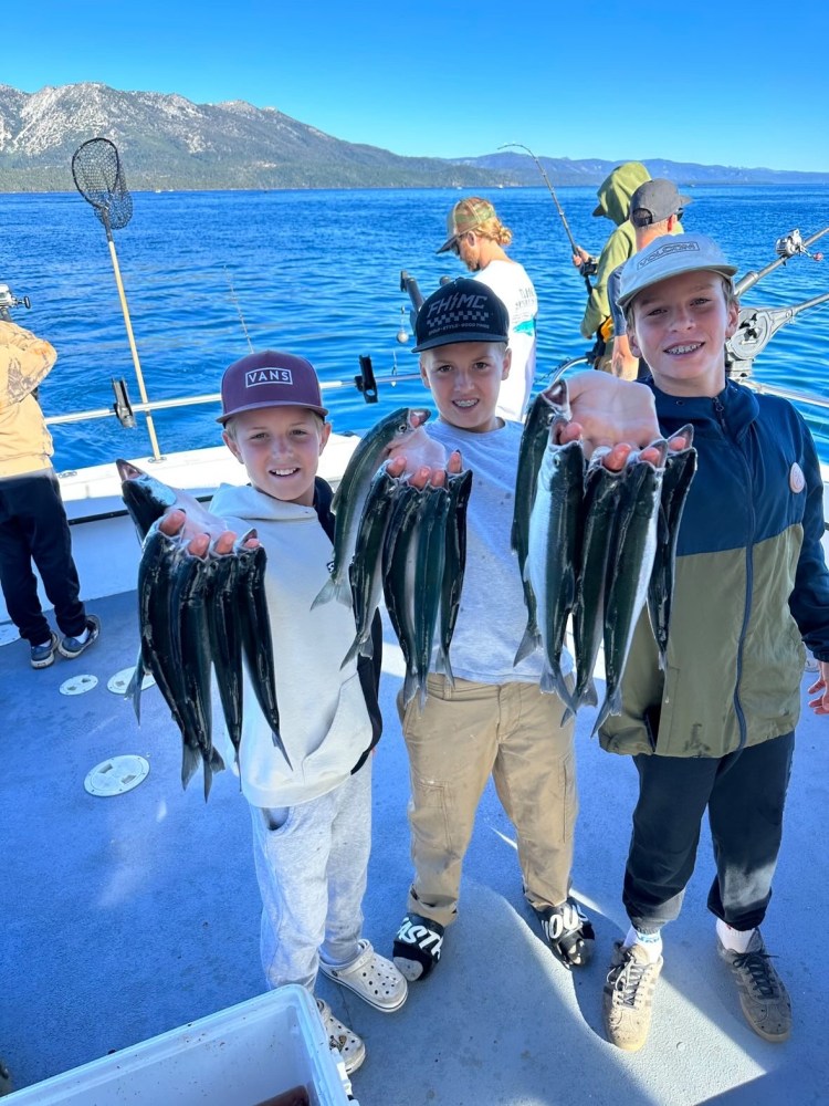 Three kids holding fish on a boat with a scenic mountain lake background.