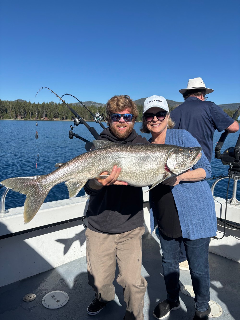 Two people holding a large fish on a boat with a lake and forest in the background.