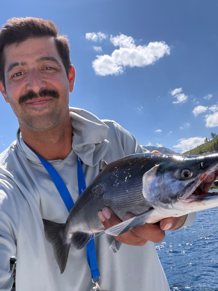 Person smiling, holding a fish against a clear blue sky with clouds.